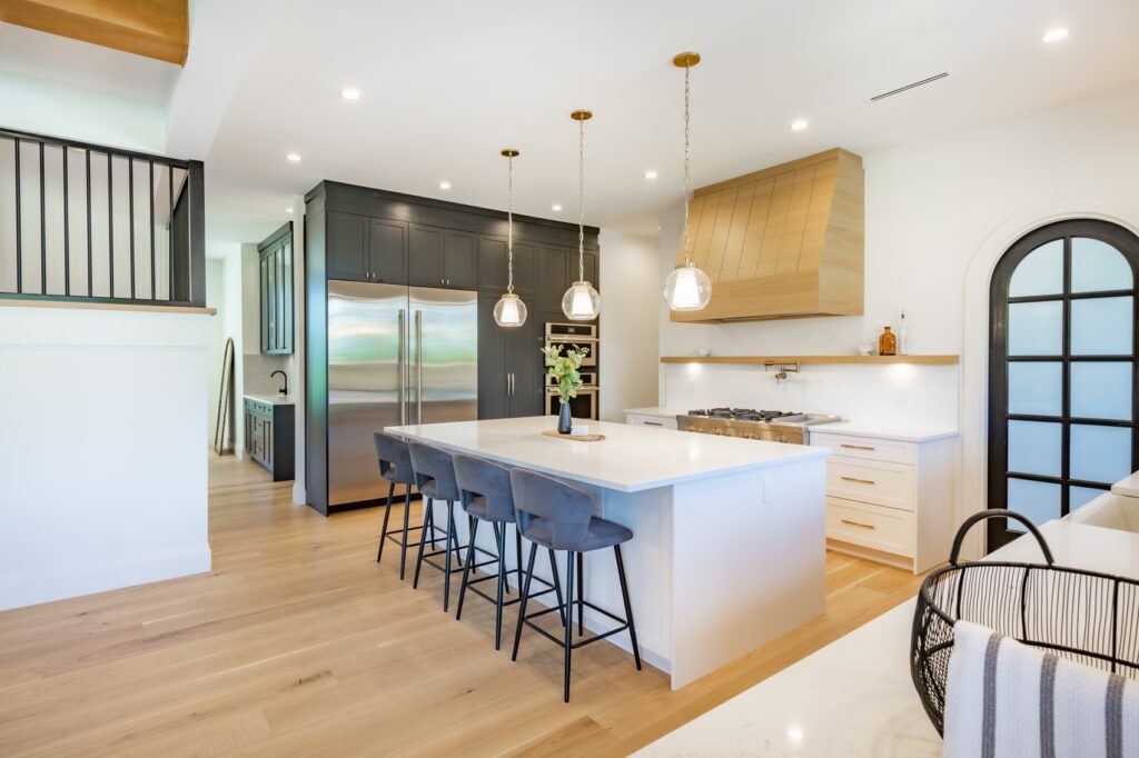 A white, modern kitchen with three hanging lights, a central island, and wood flooring.
