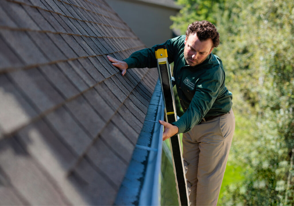 An inspector stands on a ladder and examines a gutter system.